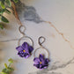 close up of the purple pansy earrings with silver findings on a marble background with foliage. 