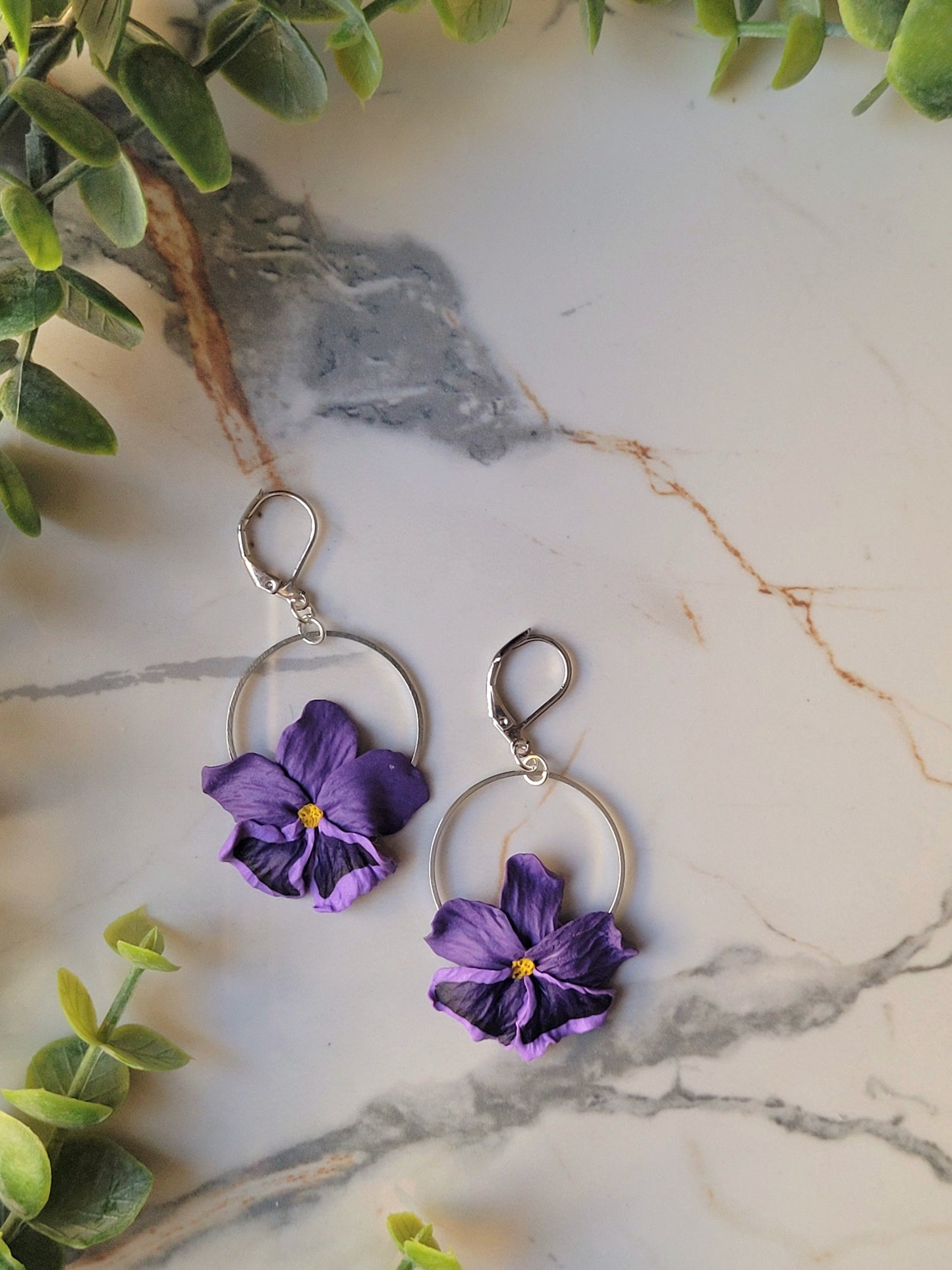 close up of the purple pansy earrings with silver findings on a marble background with foliage. 