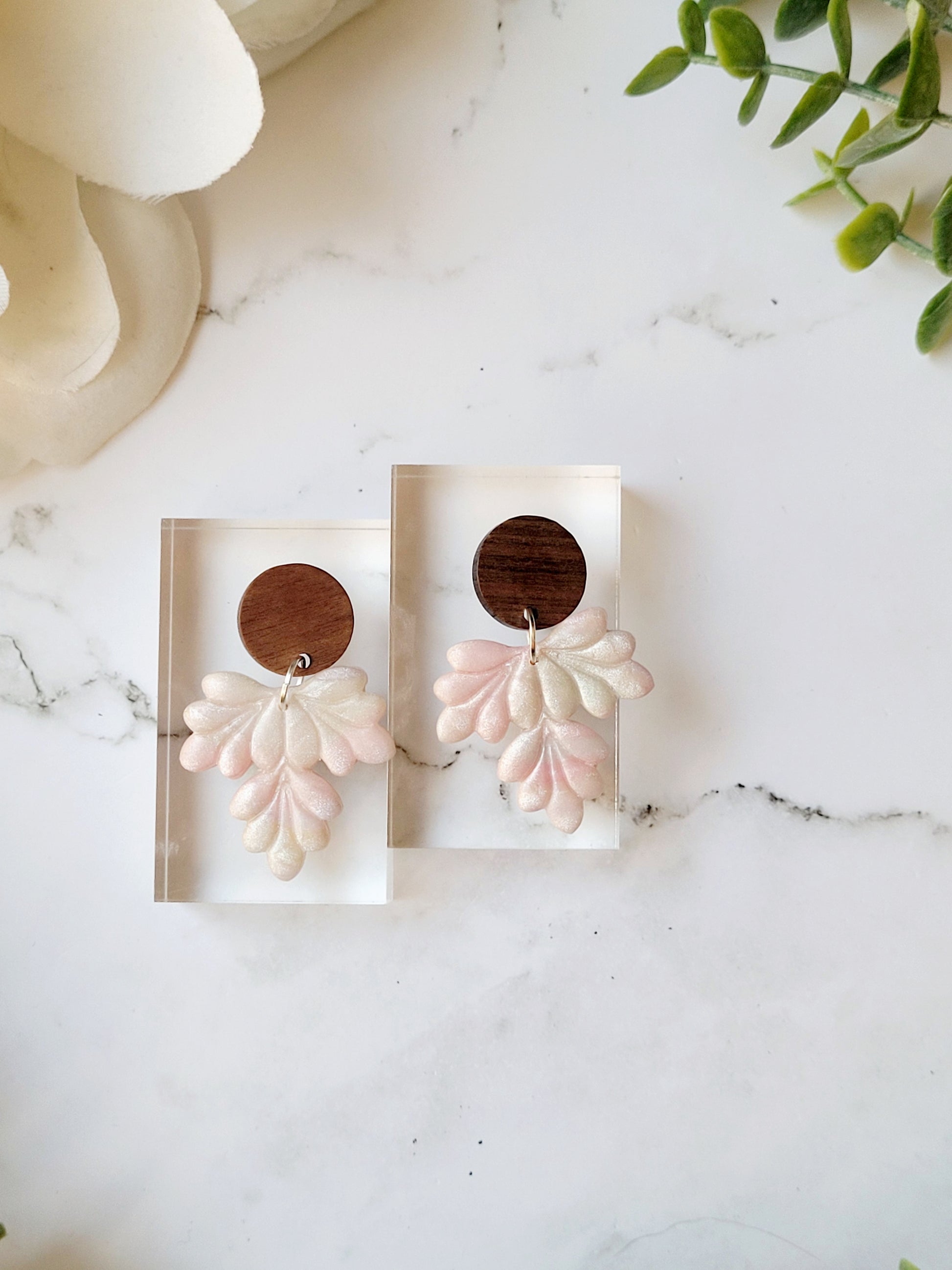 Close up of faux mother of pearl clay earrings on a marble background surrounded by foliage.