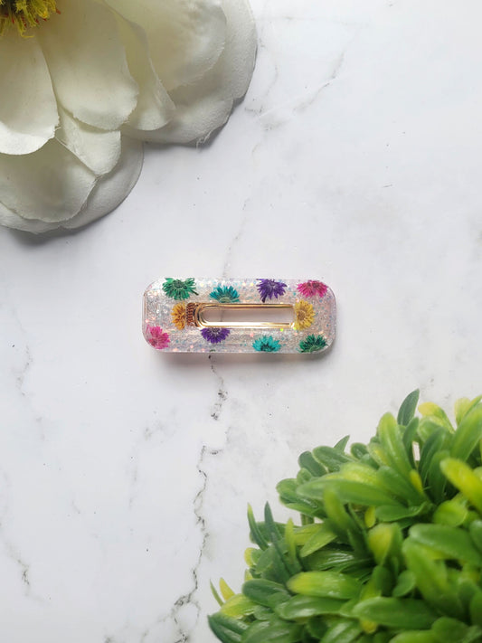close up of rectangle white glitter hair clips with rainbow dried flowers on a marble background.
