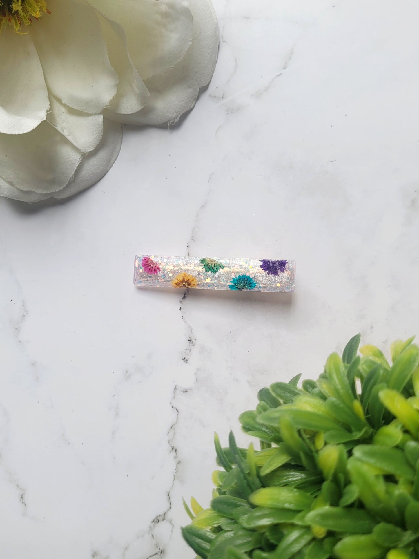 close up of slim bar white glitter hair clips with rainbow dried flowers on a marble background.