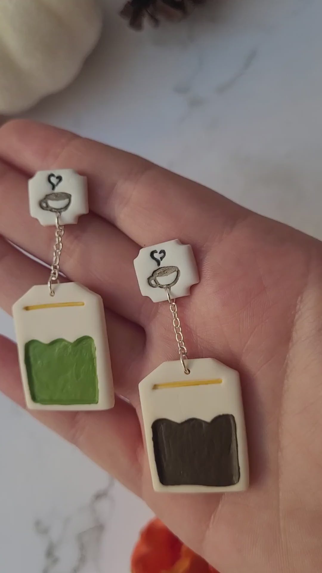 video close up of green and black tea bag shaped dangle earrings on a marble background with fall foliage.