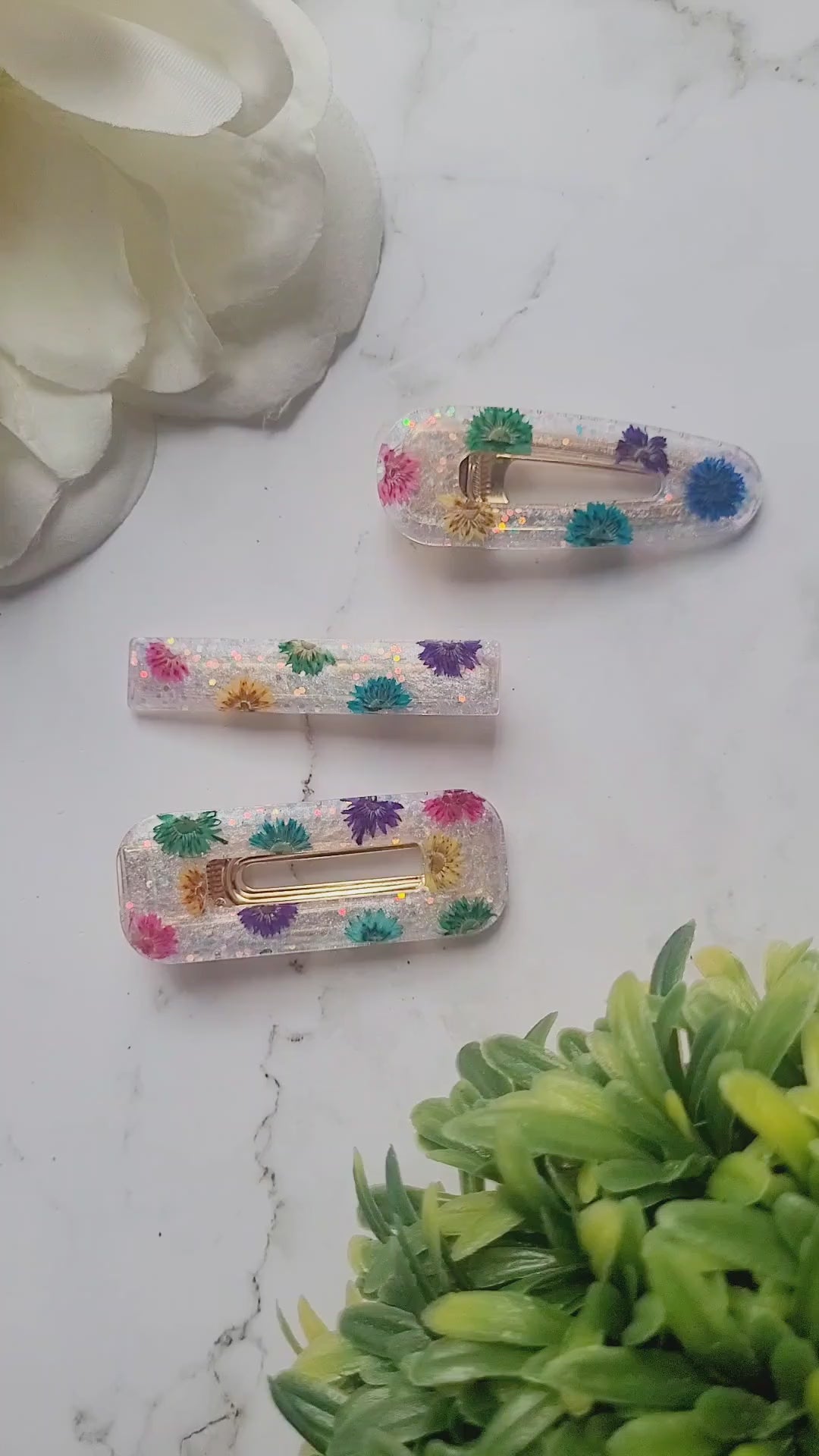 video of close up of white glitter hair clips with rainbow dried flowers on a marble background.