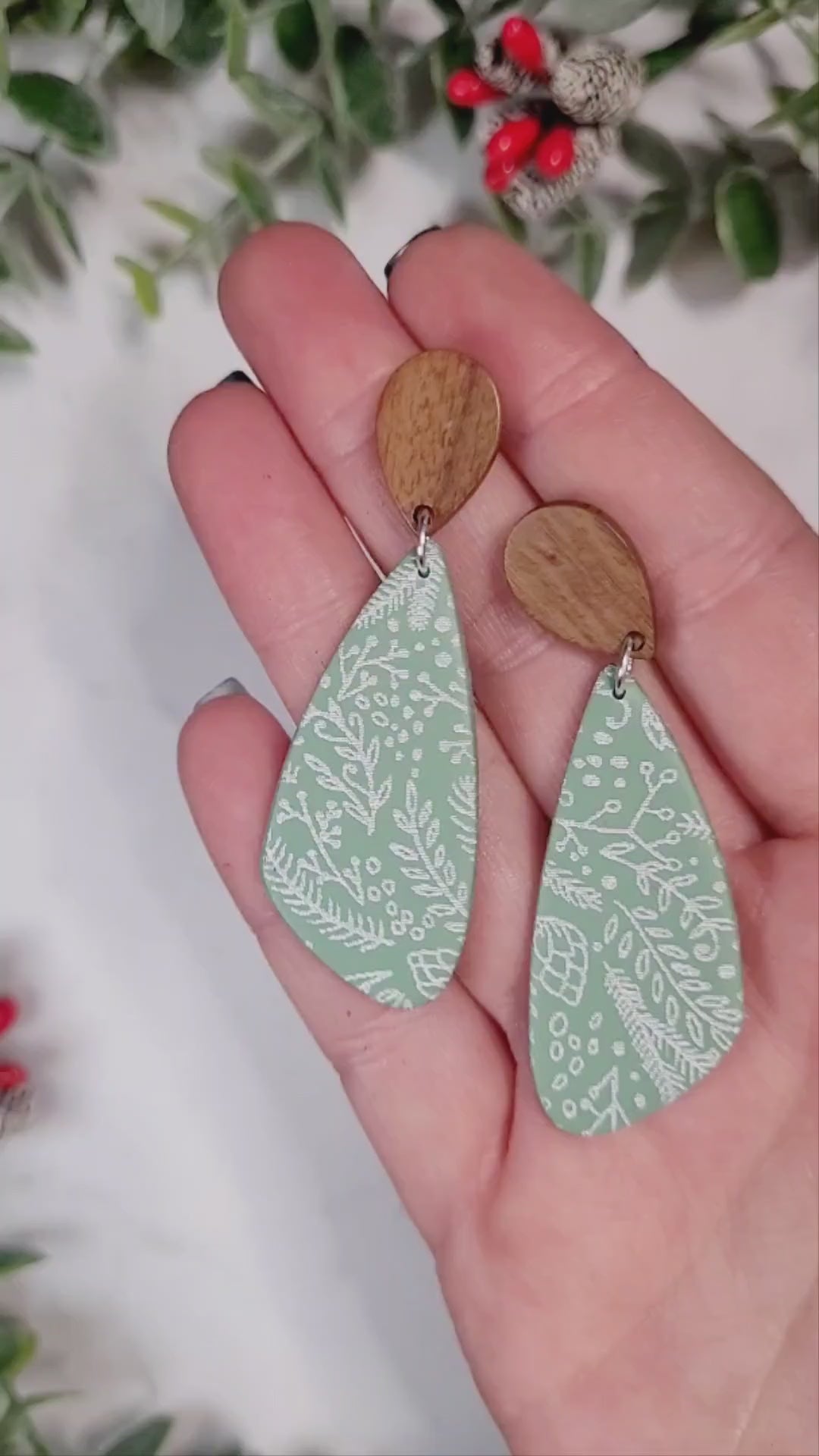 video close up of mint green tear drop earrings with a winter botanical print and wood studs on a marble background with foliage.