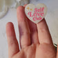 video close up of white pearl heart pin with self love club in pink on a marble background. 