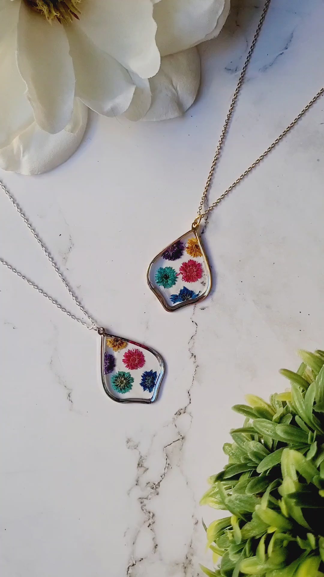 video close up of scalloped resin necklace with rainbow dried flowers on a marble background surrounded by foliage.