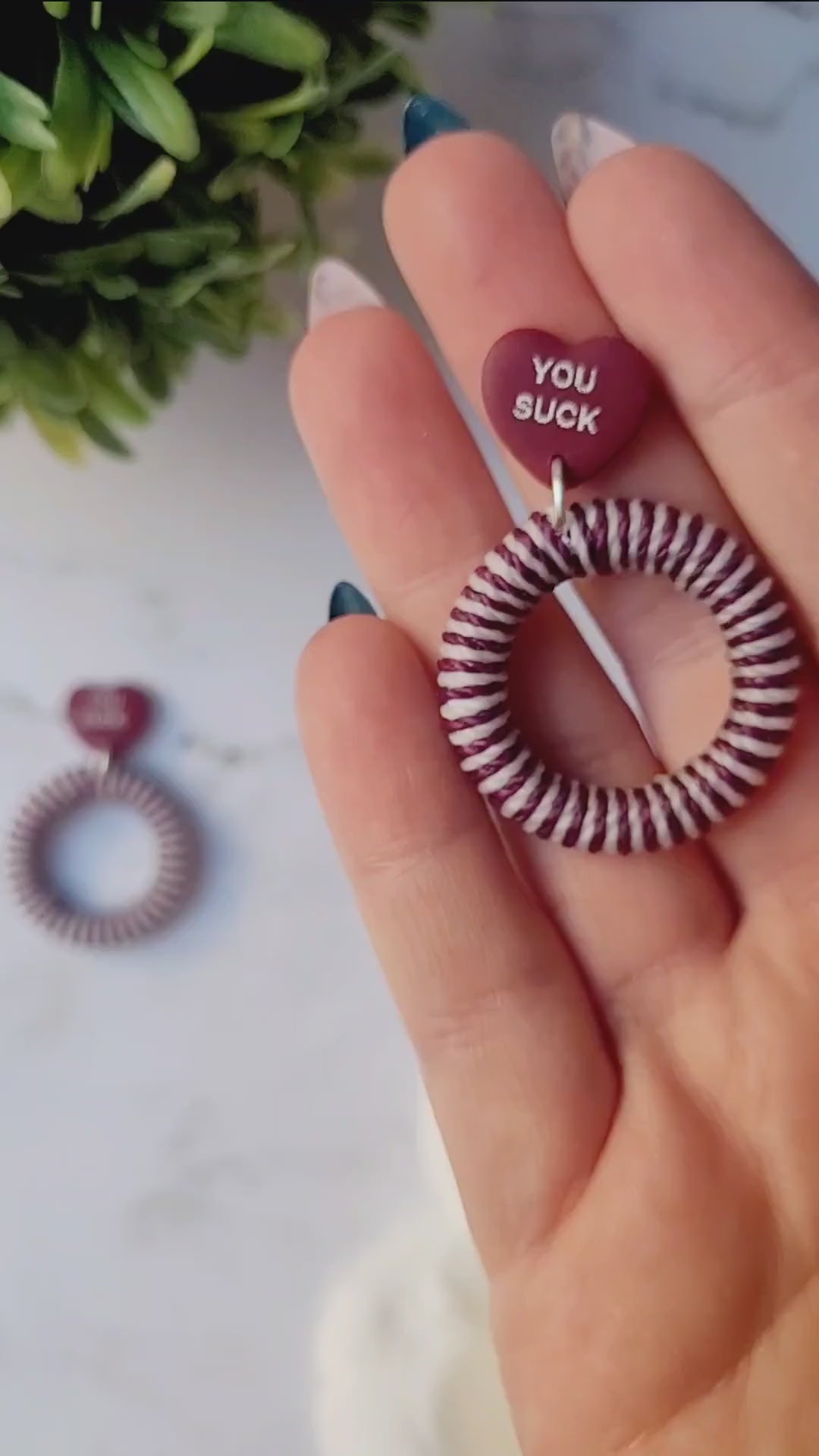 video close up of burgundy and white striped hoop with a burgundy candy heart stud on a marble background with foliage. 