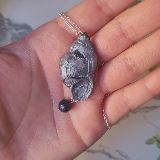 video close up of Dark silver conch shell with a black pearl necklace on a marble background surrounded by foliage.