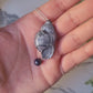 video close up of Dark silver conch shell with a black pearl necklace on a marble background surrounded by foliage.