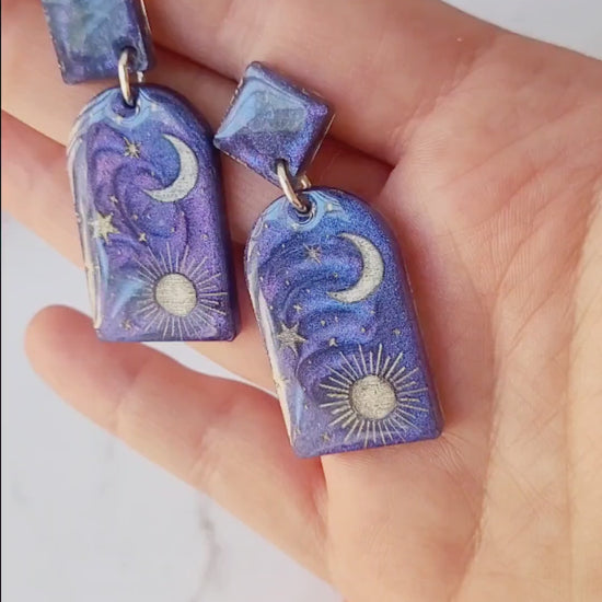 video close up of blue arch earrings with painted celestial details on a marble background.