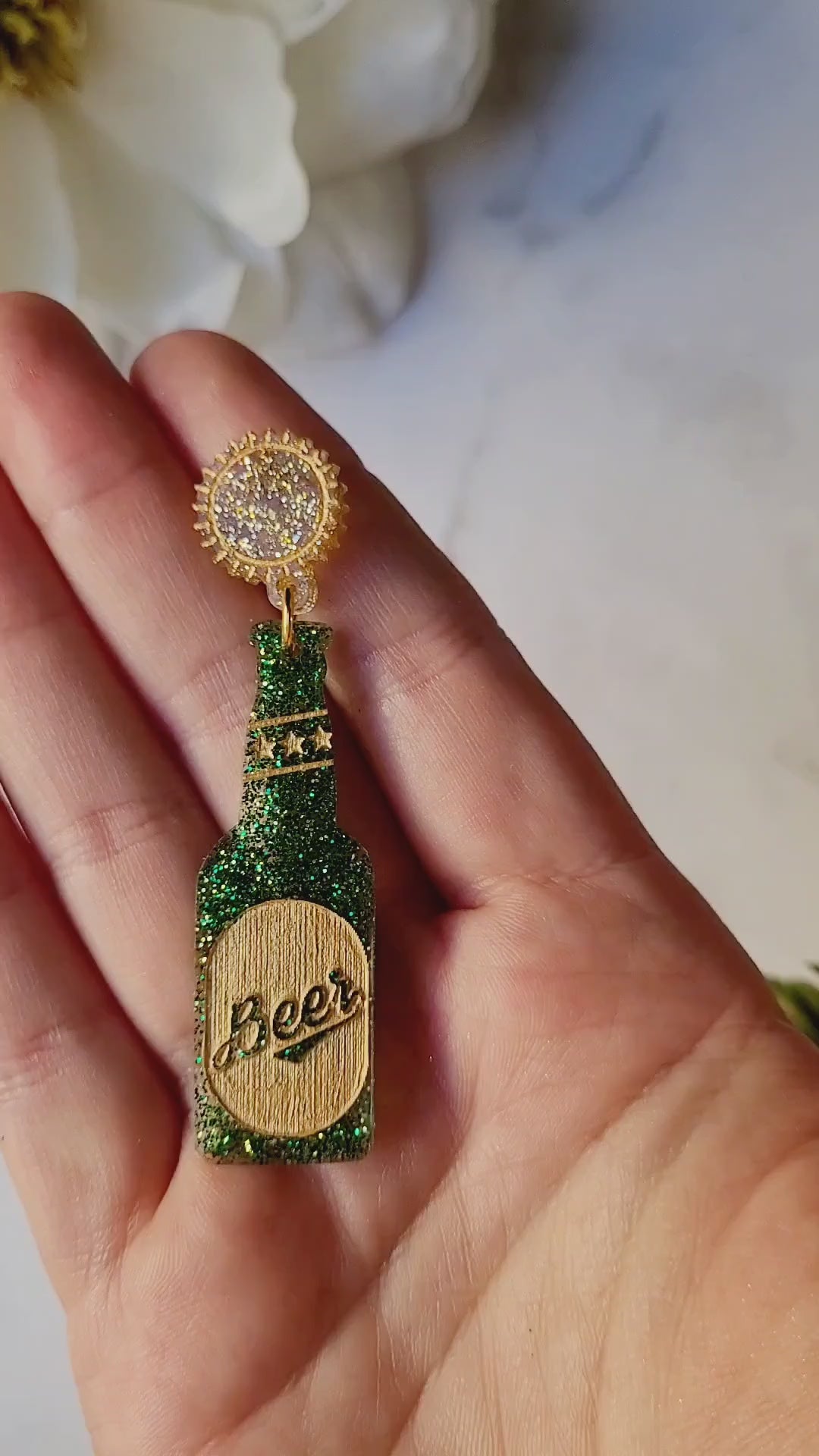 video close up of Glitter beer bottle shaped earrings on a white marble background.