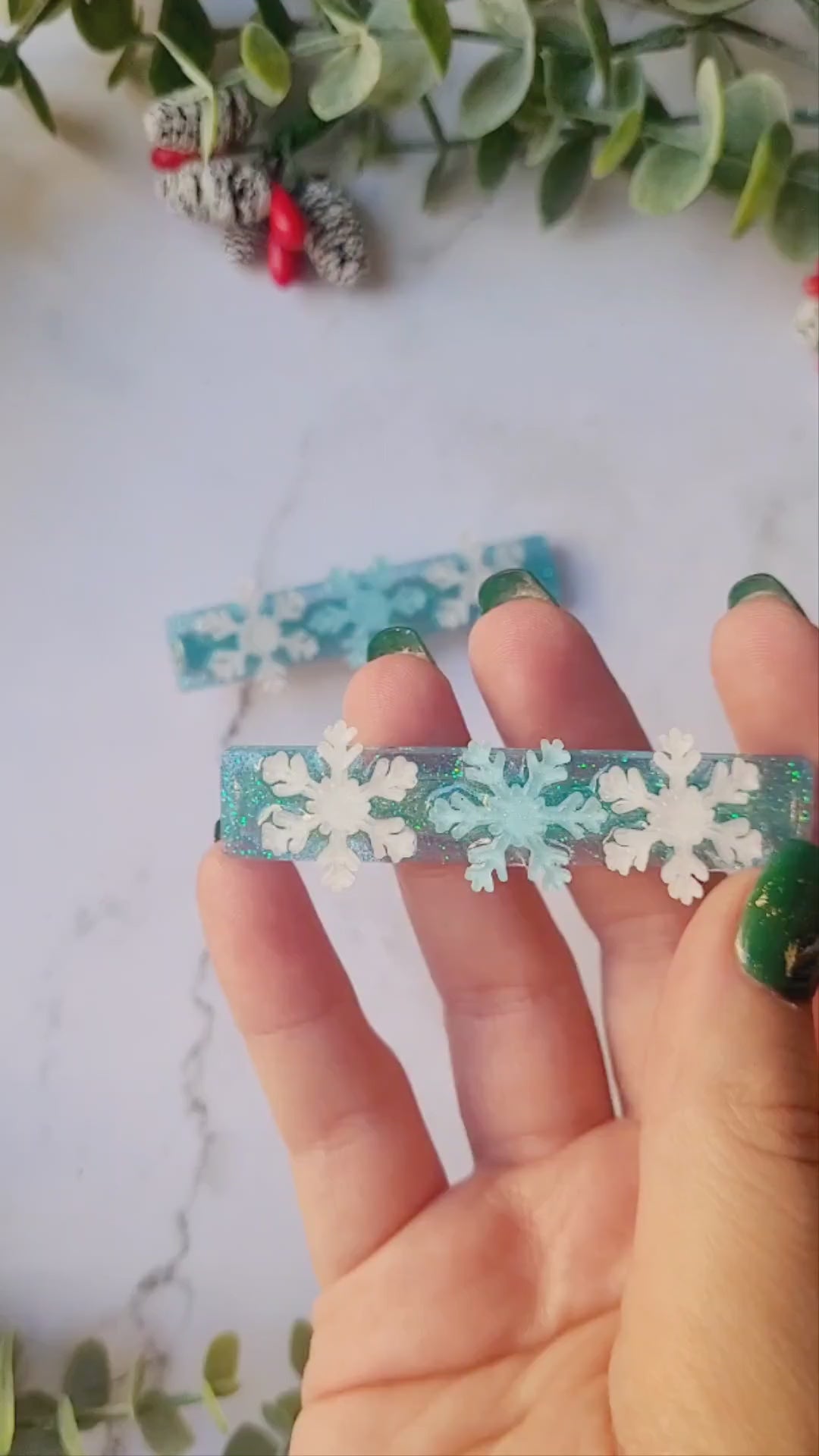 video close up of blue glitter hair clip with white and light blue snowflakes on a marble background with foliage.