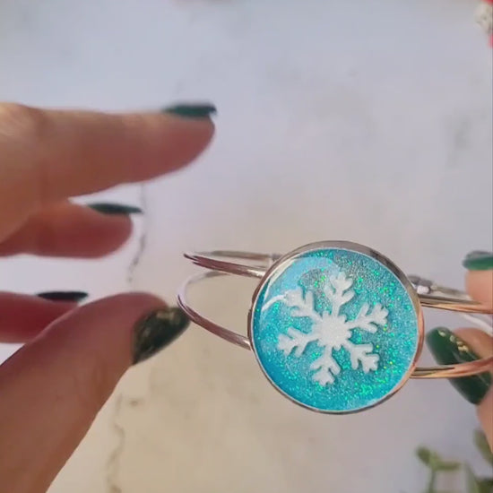 video close up of silver braceclet with a blue glitter charm and a white snowflake on white marble background with foliage.