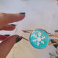 video close up of silver braceclet with a blue glitter charm and a white snowflake on white marble background with foliage.