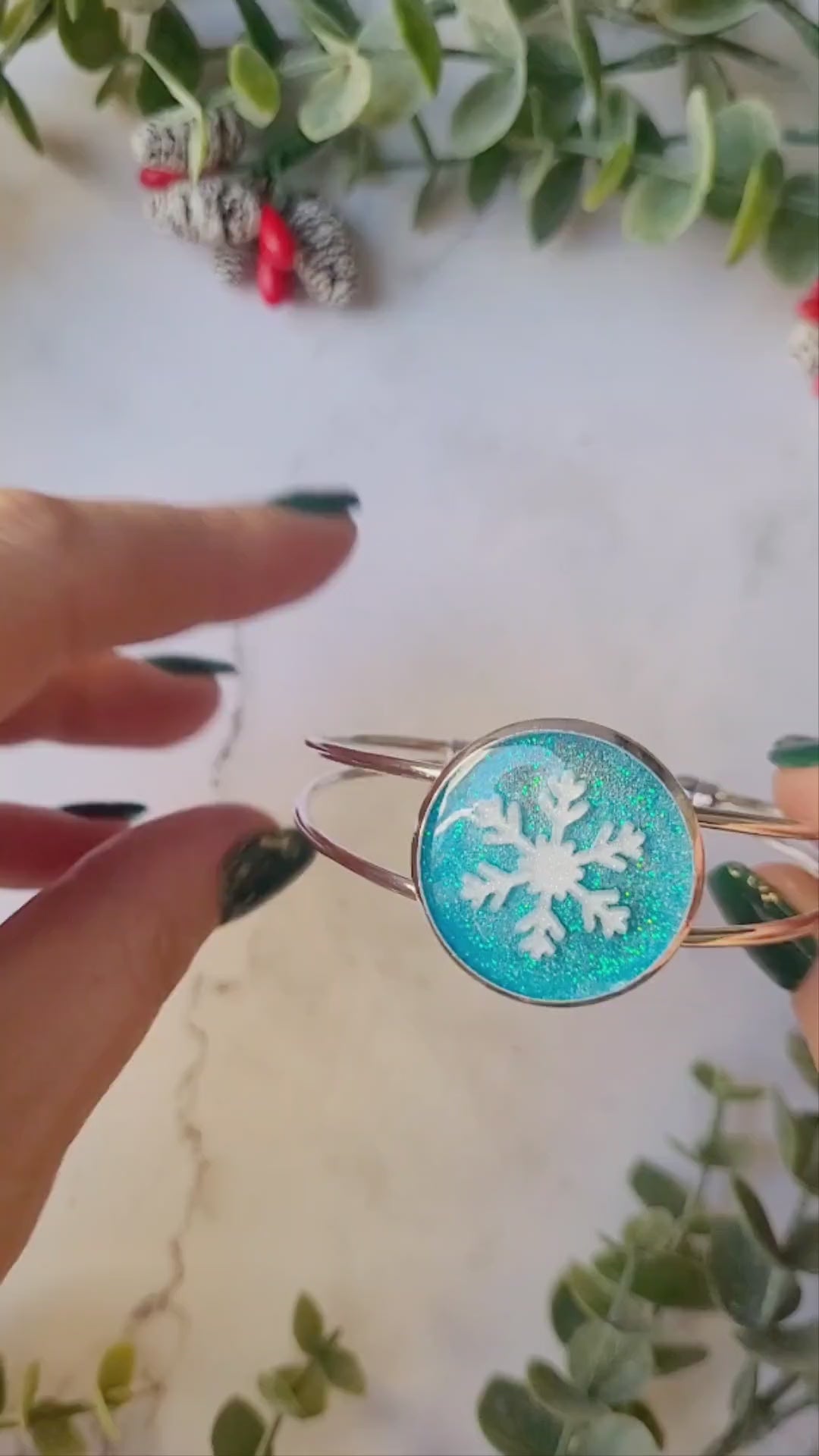 video close up of silver braceclet with a blue glitter charm and a white snowflake on white marble background with foliage.