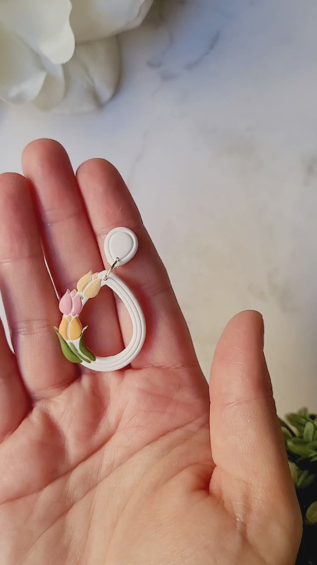 video close up of white teardrop hoops with painted tulips on a marble background with foliage.