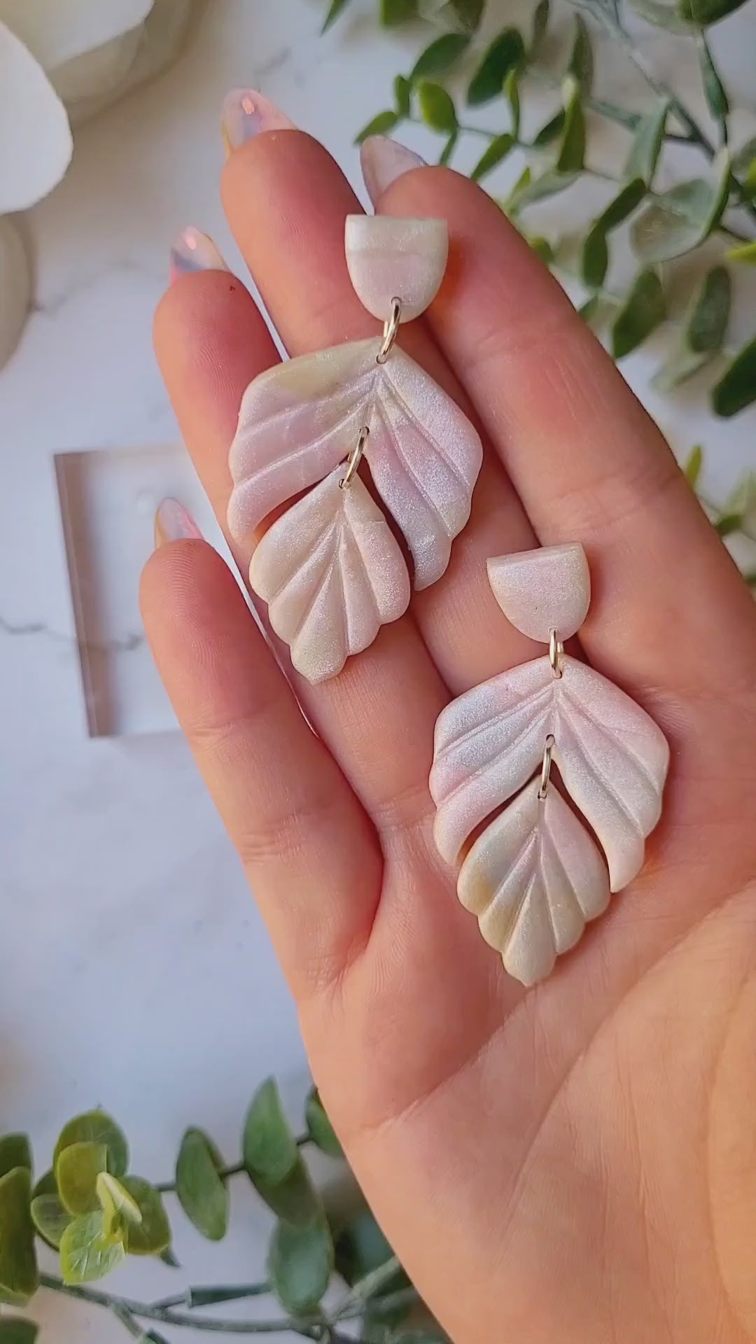 video close up of faux mother of pearl clay earrings on a marble background surrounded by foliage.