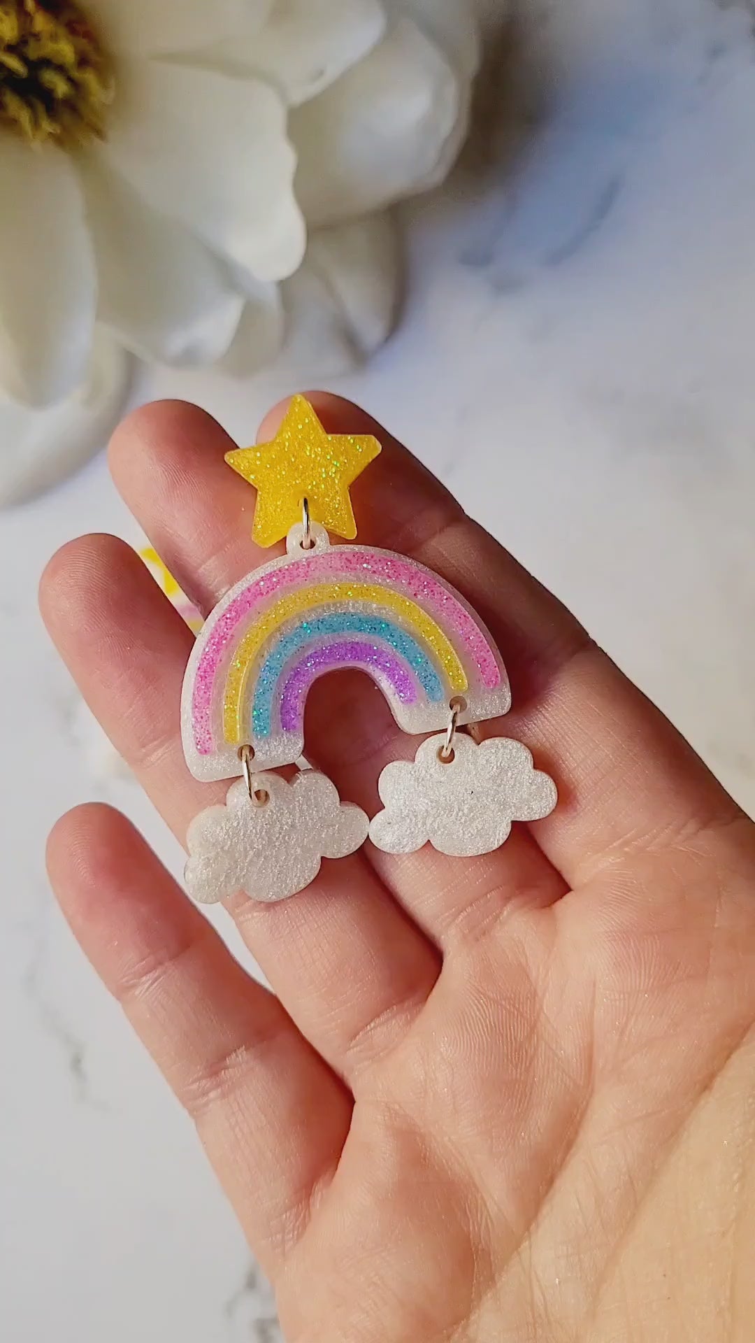 video close up of Rainbow dangle earrings with a star stud and cloud charms on a marble background with foliage.
