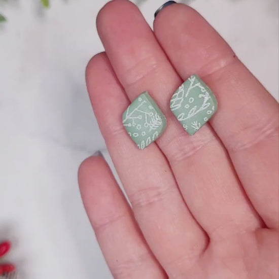 video close up of mint green diamond shaped studs with a white botanical design on a marble background with foliage. 