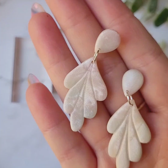 video close up of faux mother of pearl clay earrings on a marble background surrounded by foliage.