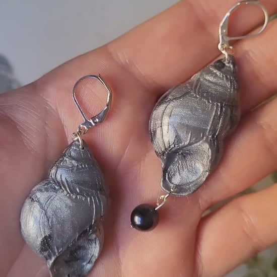 video close up of Dark silver conch shell with a black pearl on a marble background surrounded by foliage.