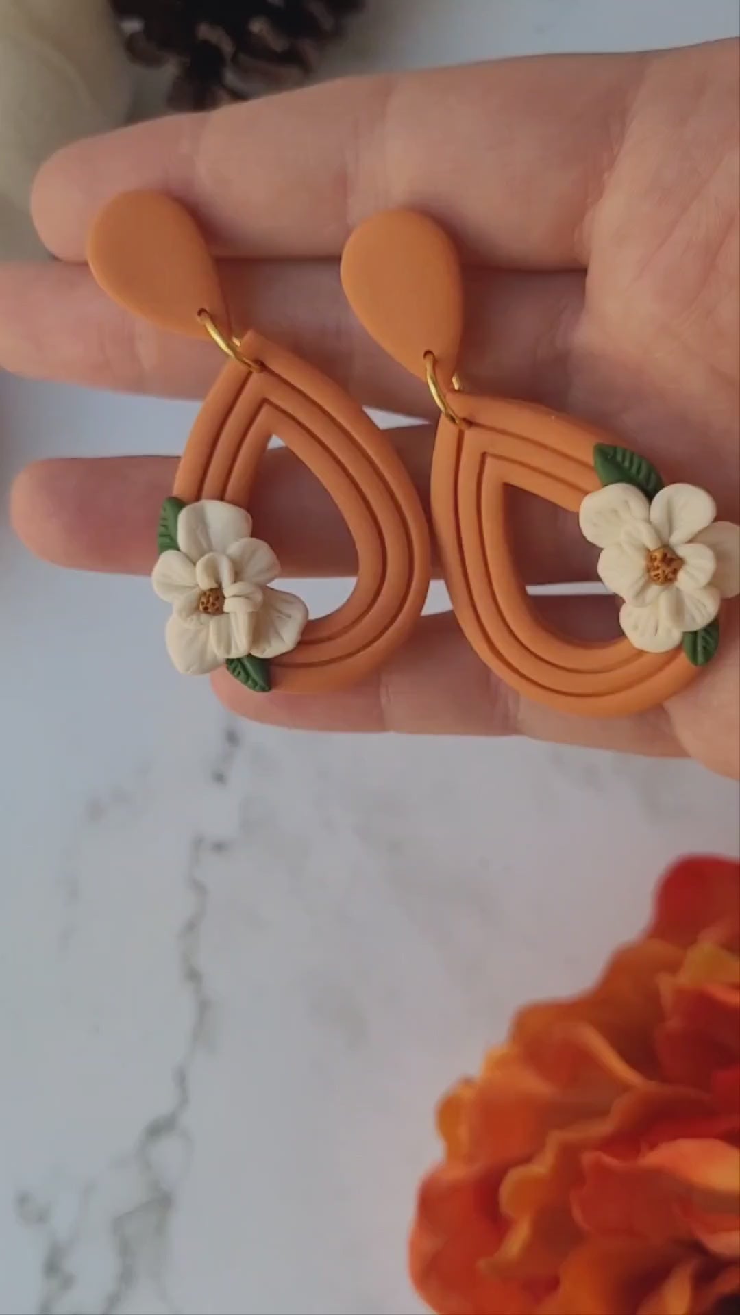 video close up of orange teardrop earrings with a white magnolia flower sitting on a marble background.