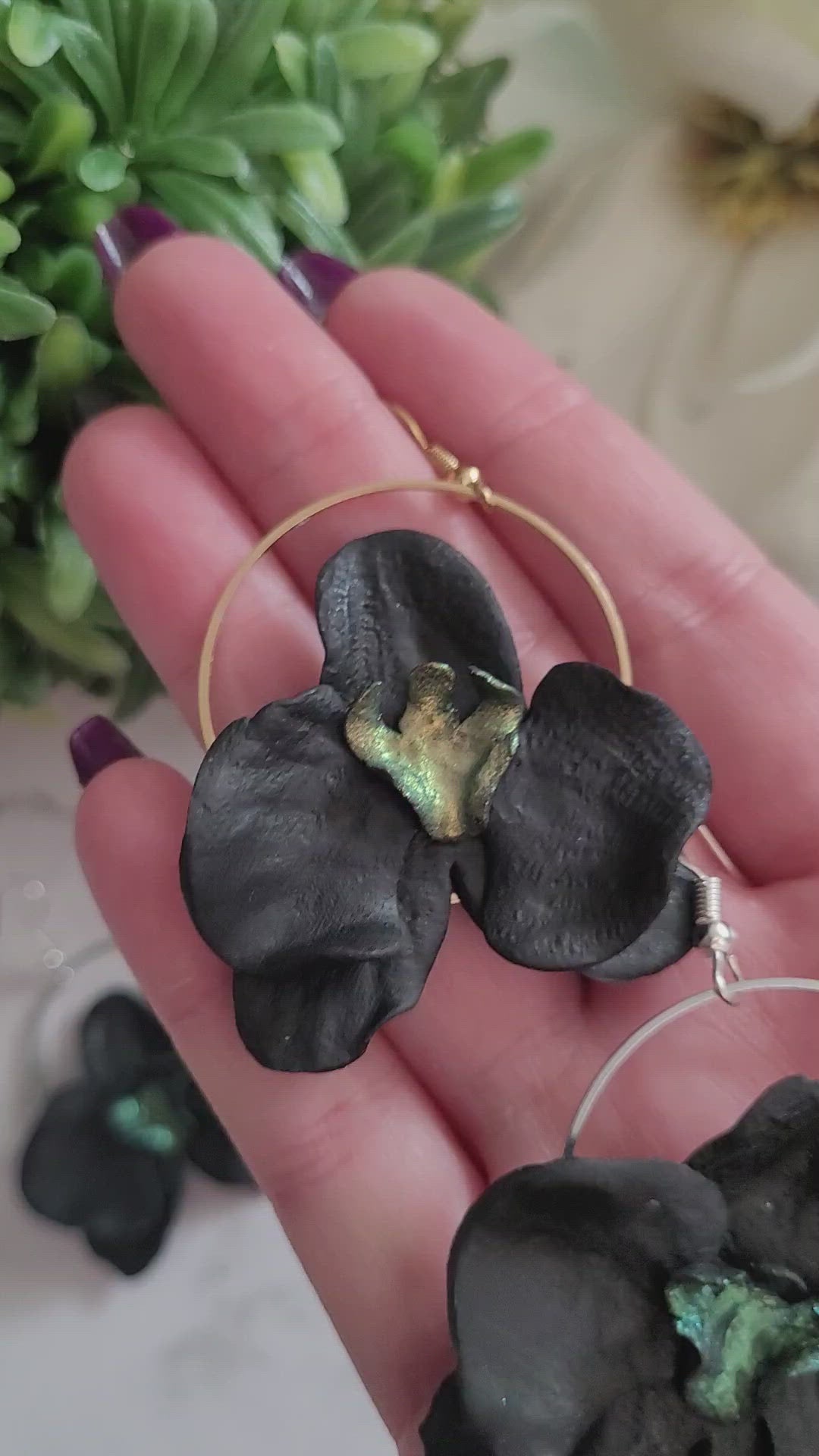 Closeup of three color variations in palm of hand. Earrings are made of black polymer clay in the shape of an orchid. 