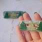 video close up of Green glitter hair clips with christmas tree charms on a white marble background with foliage.