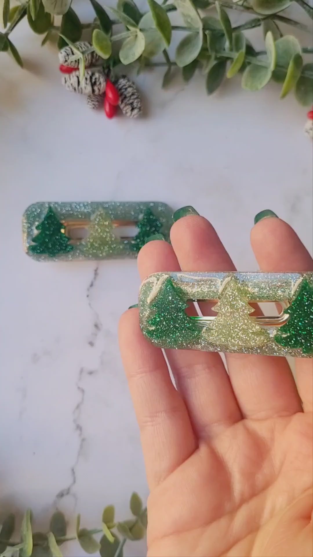 video close up of Green glitter hair clips with christmas tree charms on a white marble background with foliage.